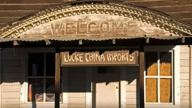 Imagem da fachada de uma pequena loja, com uma porta no meio, duas janelas de cada lado. Ao centro, está escrito 'bem-vindo', e abaixo, 'importados chineses Lokce', em inglês