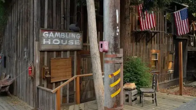 Imagem de uma casa de madeira, com a bandeira dos Estados Unidos pendurada e uma placa escrito 'casa de jogos', em inglês