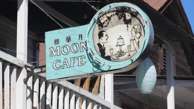 Imagem de uma placa onde se lê 'Moon Café', seguido de um desenho de um homem e uma mulher em uma mesa conversando 