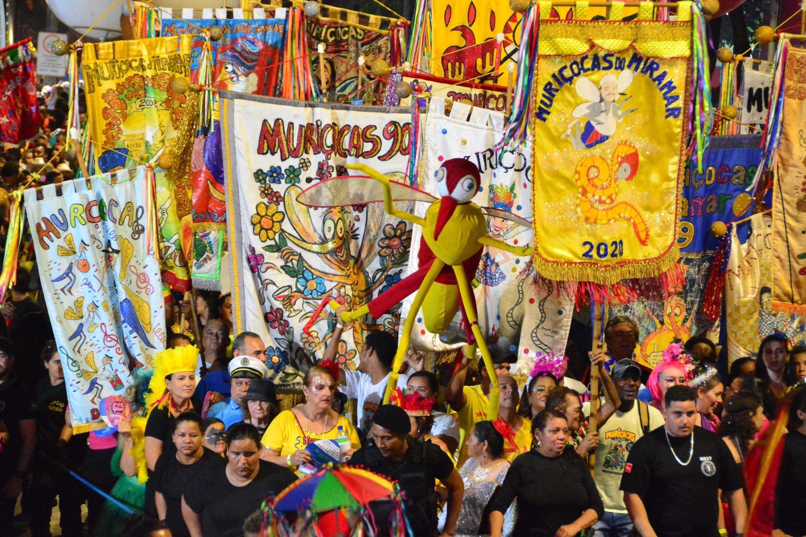 Foliões celebram Muriçocas do Miramar com estandartes coloridos e boneco gigante de muriçoca. Carnaval de João Pessoa.